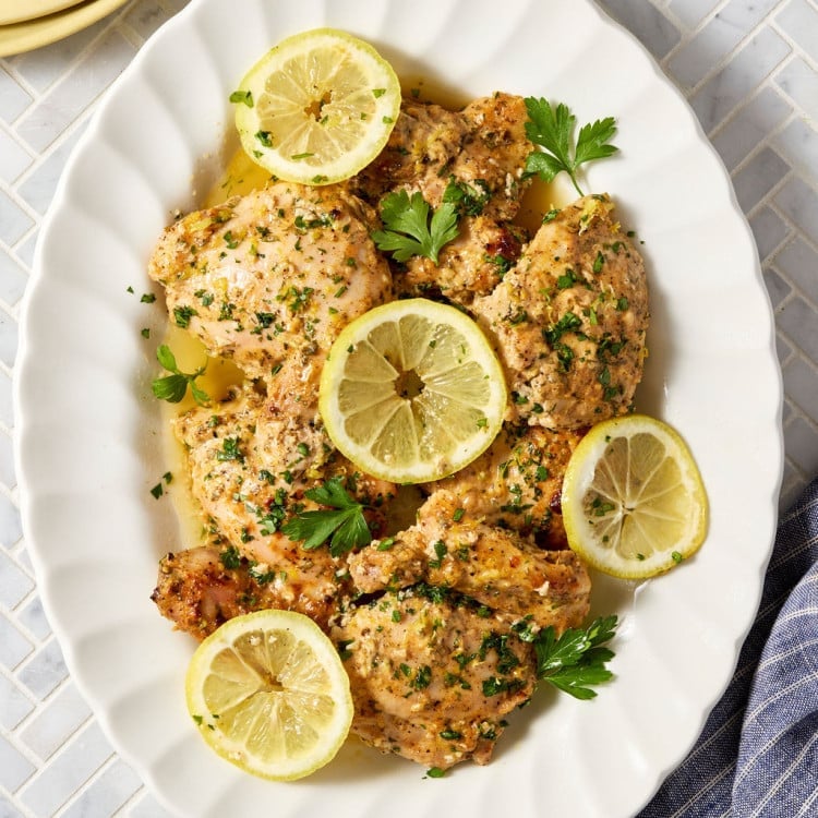 Platter of baked Greek yogurt lemon chicken topped with fresh parsley, lemon zest, and lemon slices, served with flavorful pan juices spooned over the chicken.