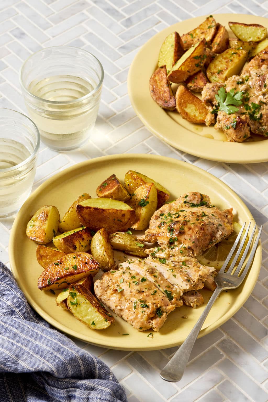 Greek yogurt lemon chicken served with roasted potatoes on a yellow plate, garnished with fresh parsley and pan juices, with glasses of white wine and a second plated serving in the background.