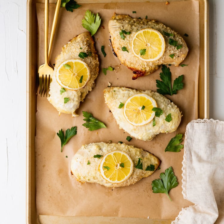 chicken breasts on a baking sheet lined with parchment paper topped with lemon slices and fresh parsley