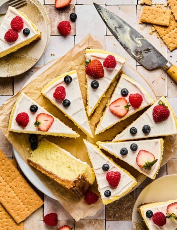 Full cheesecake topped with vanilla frosting cut into pieces, topped with berries.