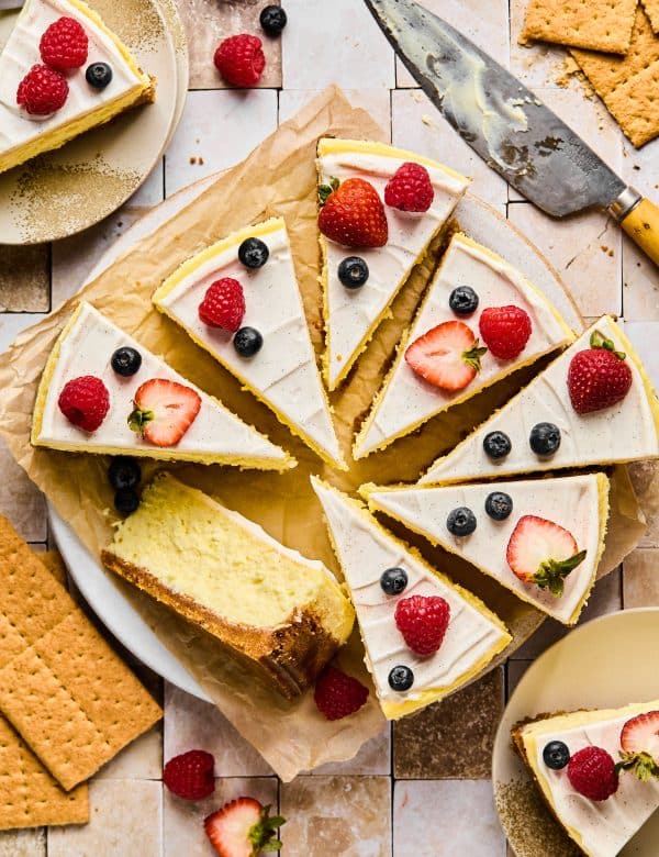 Full cheesecake topped with vanilla frosting cut into pieces, topped with berries.