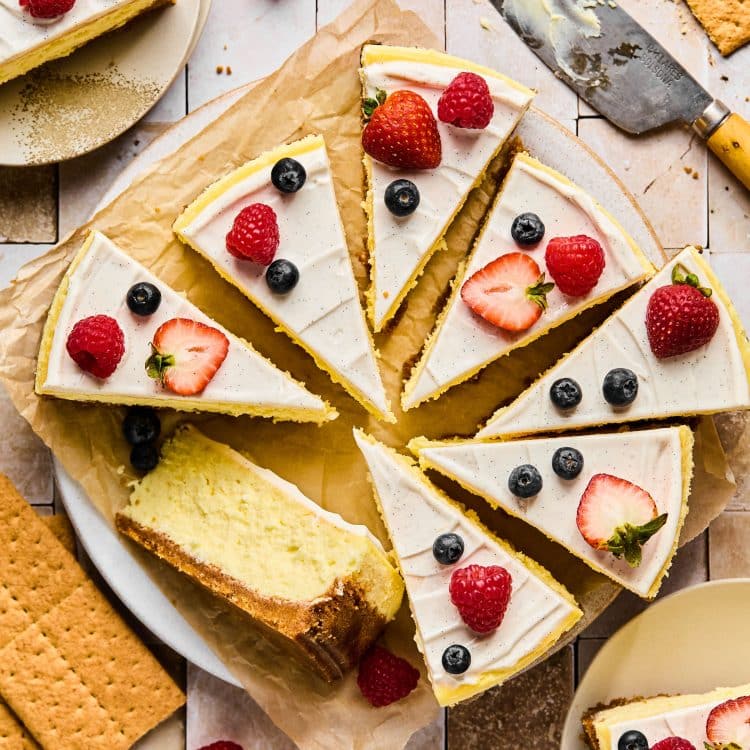 Full cheesecake topped with vanilla frosting cut into pieces, topped with berries.