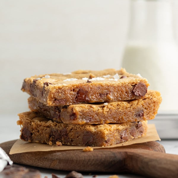 Side view of three stacked blondie bars on a wooden board with parchment paper. The blondies are thick, golden, and studded with large chocolate chunks, with a bottle of milk in the soft-focus background.
