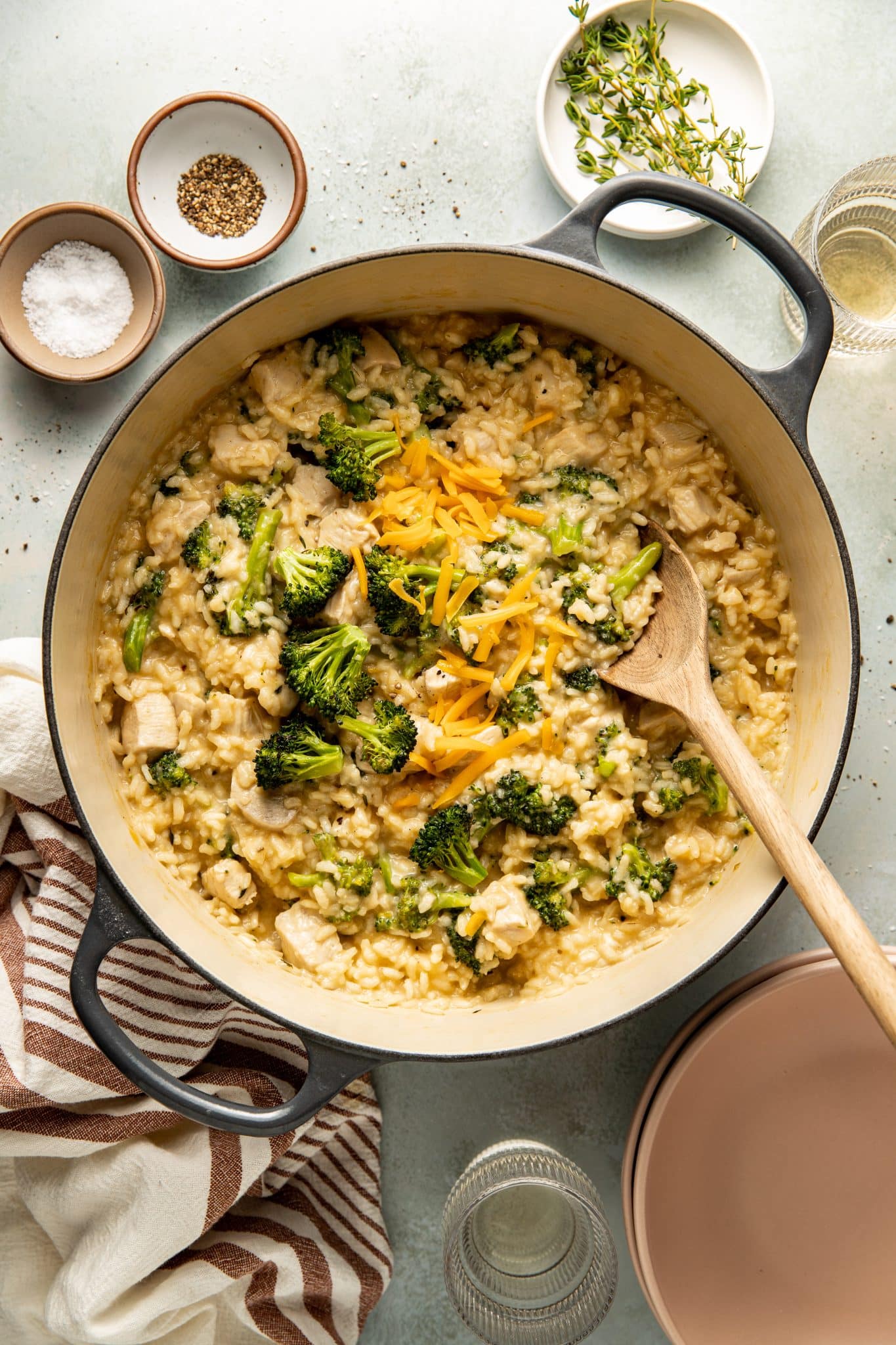 One-pot creamy chicken and broccoli risotto in a Dutch oven, garnished with shredded cheddar cheese, with a wooden spoon resting inside. Surrounded by bowls of salt, pepper, fresh thyme.