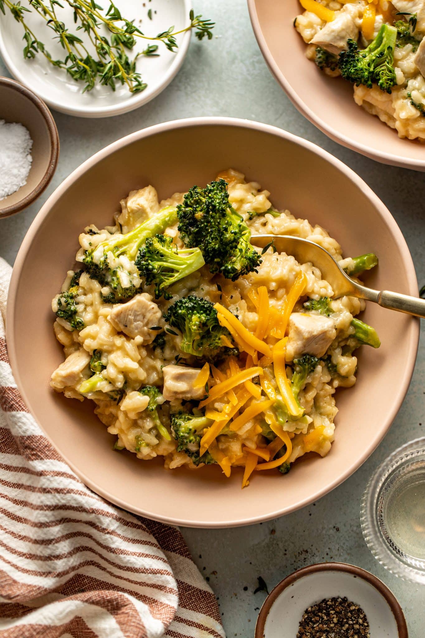 Overhead shot of a bowl of creamy chicken and broccoli risotto, topped with shredded cheddar cheese and fresh thyme. A gold spoon is partially dipped into the dish, surrounded by small bowls of salt, pepper, and fresh herbs on a rustic tabletop