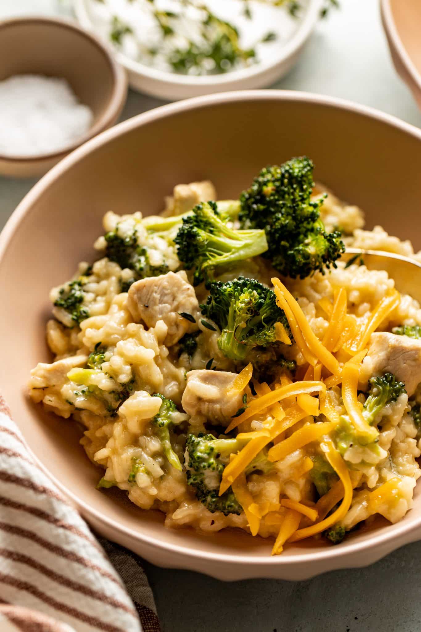 Close-up of a bowl of creamy chicken and broccoli risotto, garnished with shredded cheddar cheese and fresh thyme. The dish is served in a neutral-toned bowl with a cozy striped napkin and small bowls of salt and herbs in the background.