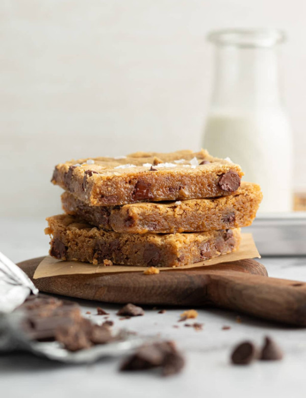 Side view of three stacked blondie bars on a wooden board with parchment paper. The blondies are thick, golden, and studded with large chocolate chunks, with a bottle of milk in the soft-focus background.