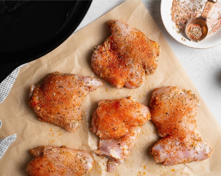 Raw boneless, skinless chicken thighs seasoned with Italian seasoning, paprika, garlic powder, salt, and pepper on parchment paper before cooking.