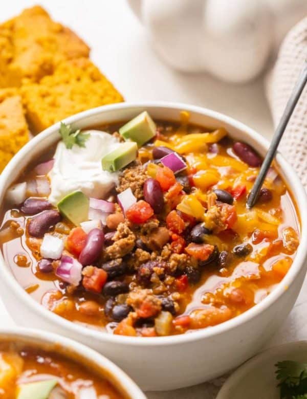 a white bowl filled with chili made with beans and garnished with red onion, cheese, sour cream and avocado.