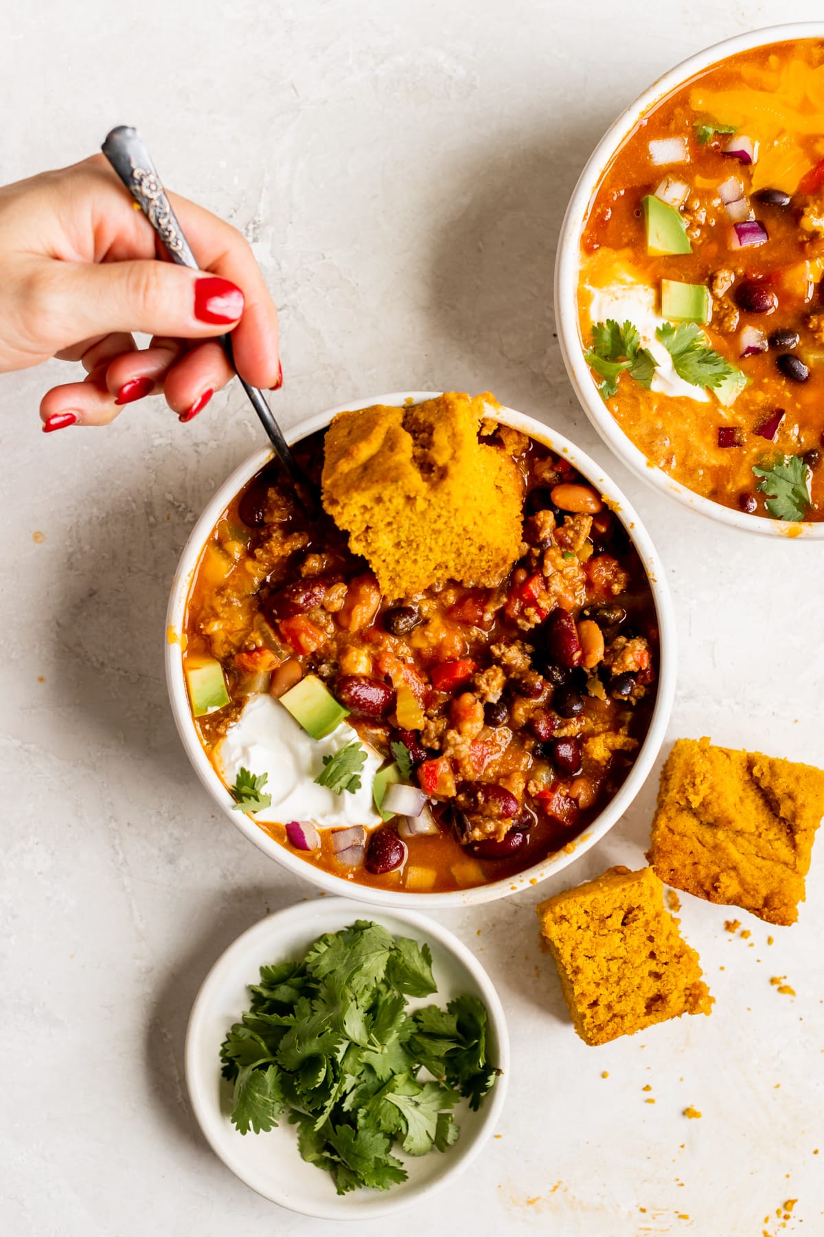 a white bowl filled with chili made with beans and garnished with red onion, cheese, cilantro, cornbread sour cream and avocado. 