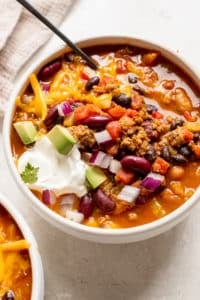 a white bowl filled with chili made with beans and garnished with red onion, cheese, sour cream and avocado.