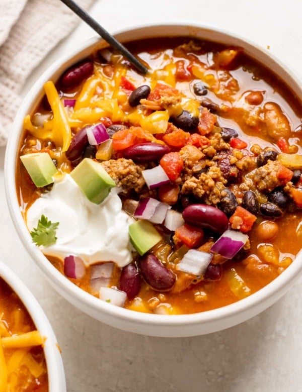 a white bowl filled with chili made with beans and garnished with red onion, cheese, sour cream and avocado.