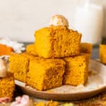 squares of pumpkin cornbread stacked on a tan plate and topped with a scoop of butter and a drizzle of honey