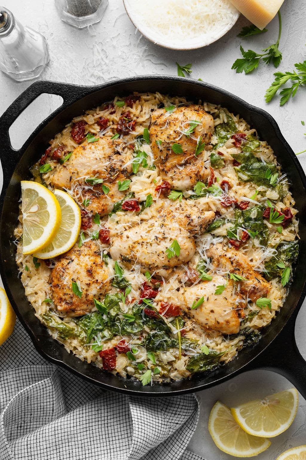 Overhead view of creamy Tuscan chicken and rice in a cast iron skillet with golden-browned chicken thighs, creamy Parmesan rice, spinach, sun-dried tomatoes, fresh parsley, grated Parmesan, and lemon slices for garnish.