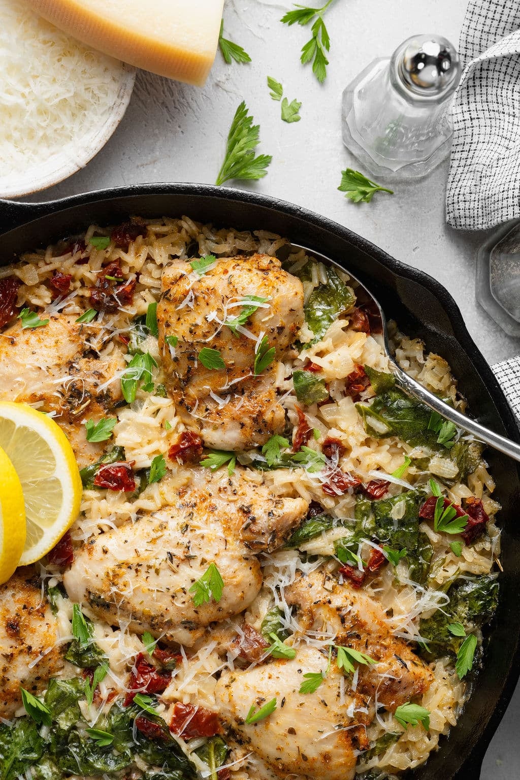 Close-up overhead view of creamy Tuscan chicken and rice in a cast iron skillet with browned chicken thighs, creamy Parmesan rice, spinach, sun-dried tomatoes, fresh parsley, grated Parmesan, and lemon slices.