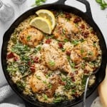 Overhead view of a cast iron skillet filled with creamy Tuscan chicken and rice, featuring golden-browned chicken thighs nestled in creamy Parmesan rice with spinach and chopped sun-dried tomatoes. Garnished with grated Parmesan, fresh parsley, and lemon slices, with additional Parmesan, sun-dried tomatoes, and seasonings styled around the skillet.