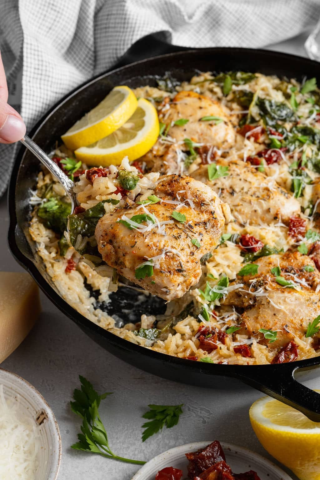 Spoon lifting a browned chicken thigh with creamy rice, spinach, and sun-dried tomatoes from a cast iron skillet of creamy Tuscan chicken and rice, garnished with Parmesan, parsley, and lemon slices.