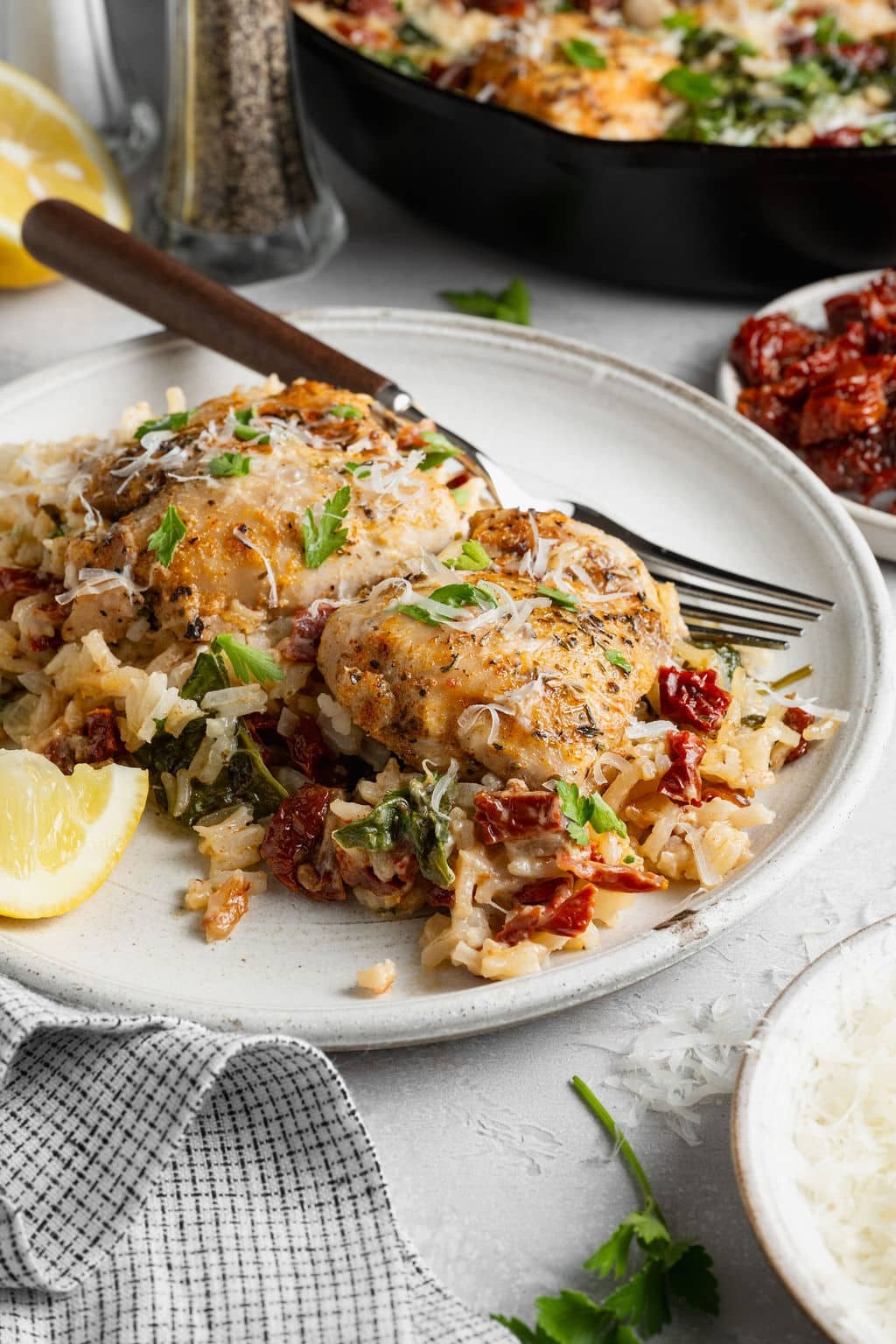 Plated serving of creamy Tuscan chicken and rice topped with browned chicken thighs, Parmesan cheese, fresh parsley, spinach, and sun-dried tomatoes, served with a lemon wedge.