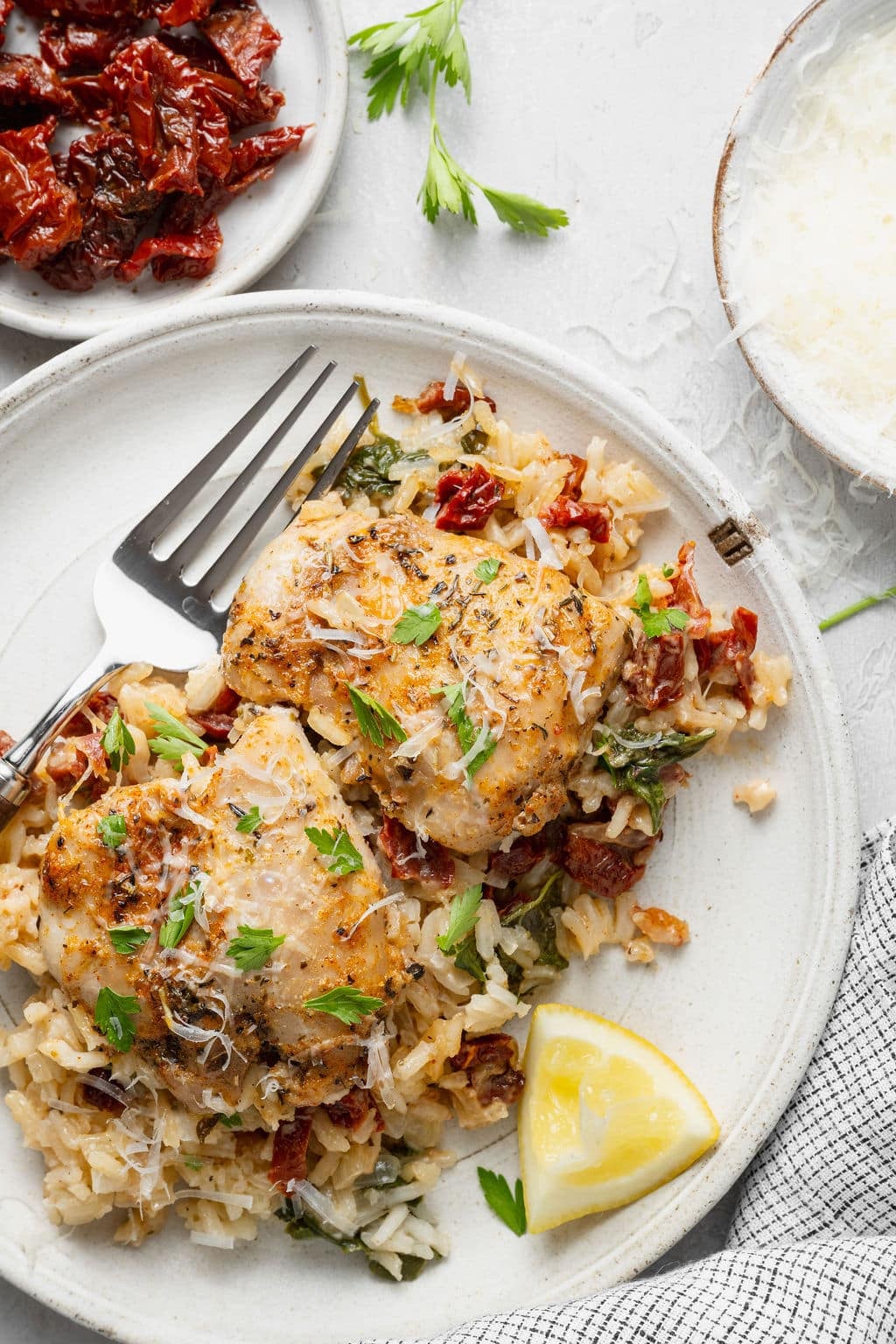 Overhead view of a plated serving of creamy Tuscan chicken and rice with browned chicken thighs, creamy Parmesan rice, spinach, sun-dried tomatoes, fresh parsley, grated Parmesan, and a lemon wedge.