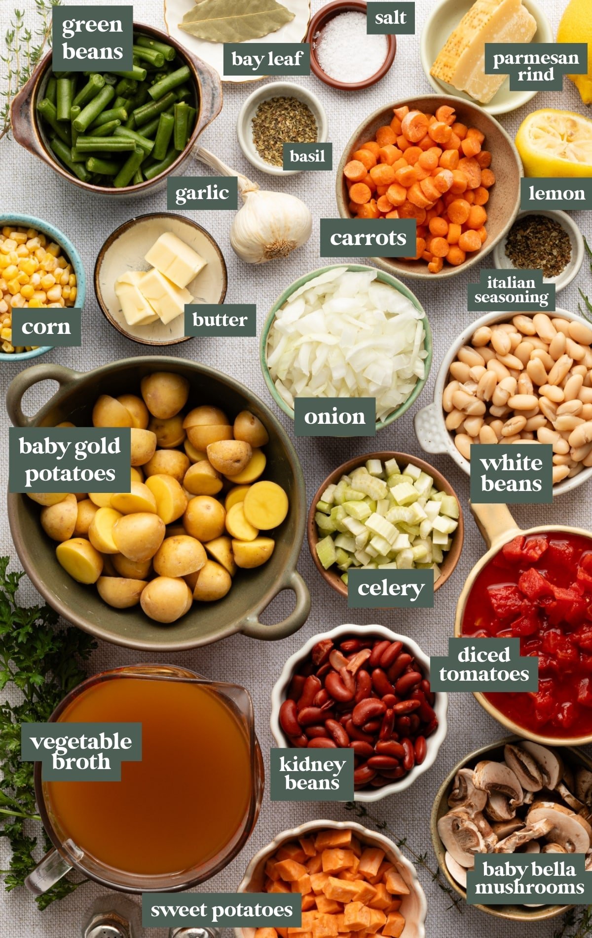 Overhead view of fresh vegetable soup ingredients arranged in bowls, including baby gold potatoes, sweet potatoes, green beans, corn, carrots, celery, onion, garlic, mushrooms, diced tomatoes, kidney beans, white beans, vegetable broth, parmesan rind, bay leaves, butter, lemon, and seasonings.