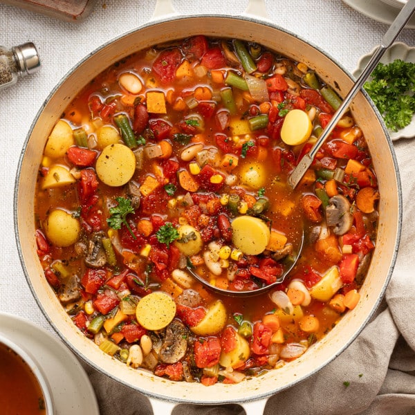 Overhead view of a Dutch oven full of vegetable soup with potatoes, carrots, beans, corn, and tomatoes, surrounded by sliced bread, fresh parsley, and serving bowls.