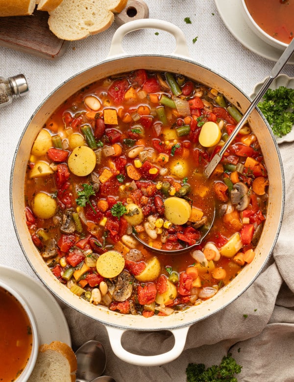 Overhead view of a Dutch oven full of vegetable soup with potatoes, carrots, beans, corn, and tomatoes, surrounded by sliced bread, fresh parsley, and serving bowls.