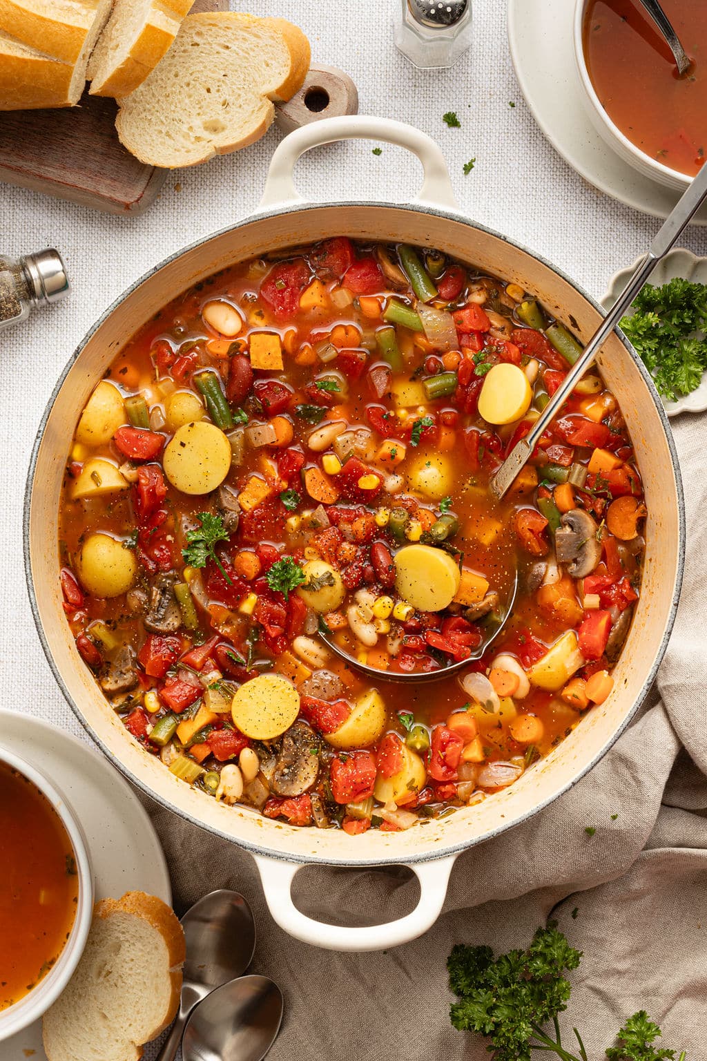 Overhead view of a Dutch oven full of vegetable soup with potatoes, carrots, beans, corn, and tomatoes, surrounded by sliced bread, fresh parsley, and serving bowls.