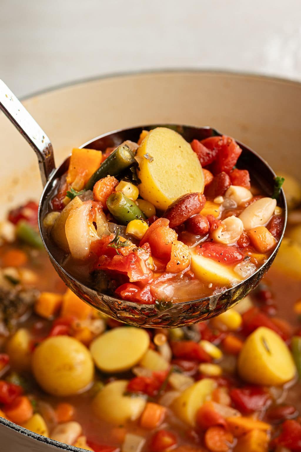 Close-up of a ladle lifting a serving of vegetable soup, showing chunks of potatoes, carrots, green beans, tomatoes, and beans in a rich broth.