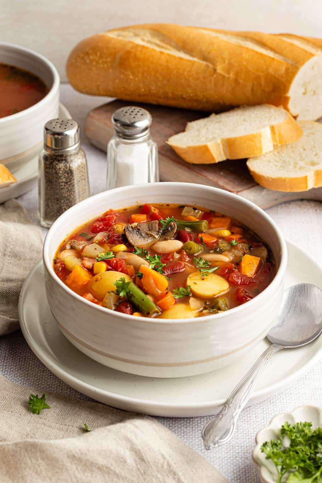 Bowl of vegetable soup on a plate with a spoon, garnished with fresh parsley, and served with slices of bread in the background.