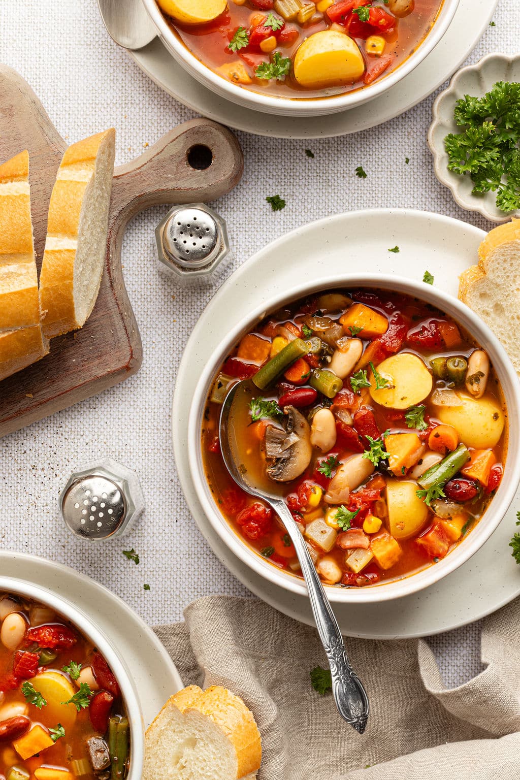 Overhead view of bowls of vegetable soup served with sliced bread, showing hearty vegetables like potatoes, carrots, mushrooms, and beans in a tomato-based broth.