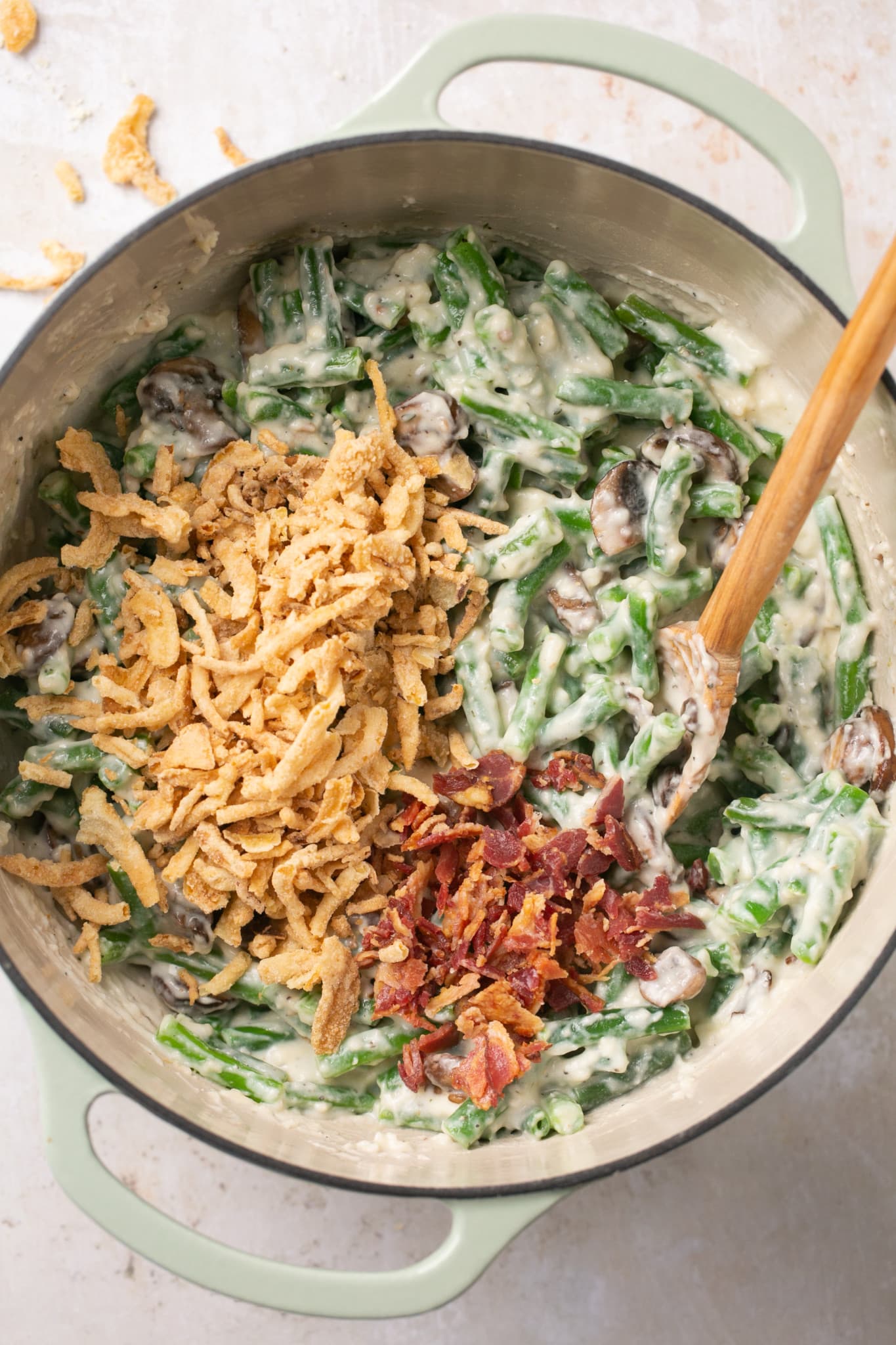 green beans mixed in a white sauce in a dutch oven with fried onions and bacon on top. 
