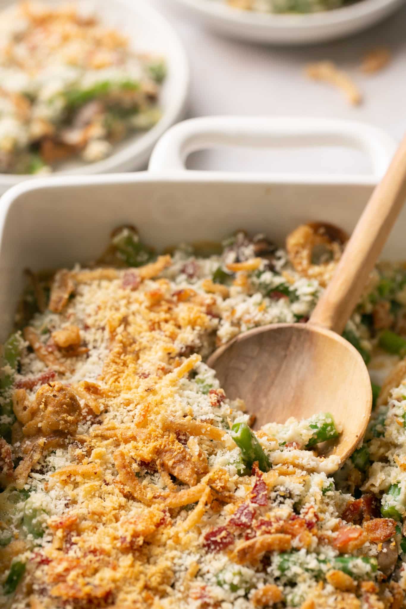 a wooden spoon in a casserole dish made with green beans and topped with panko breadcrumbs and bacon crumbles. 