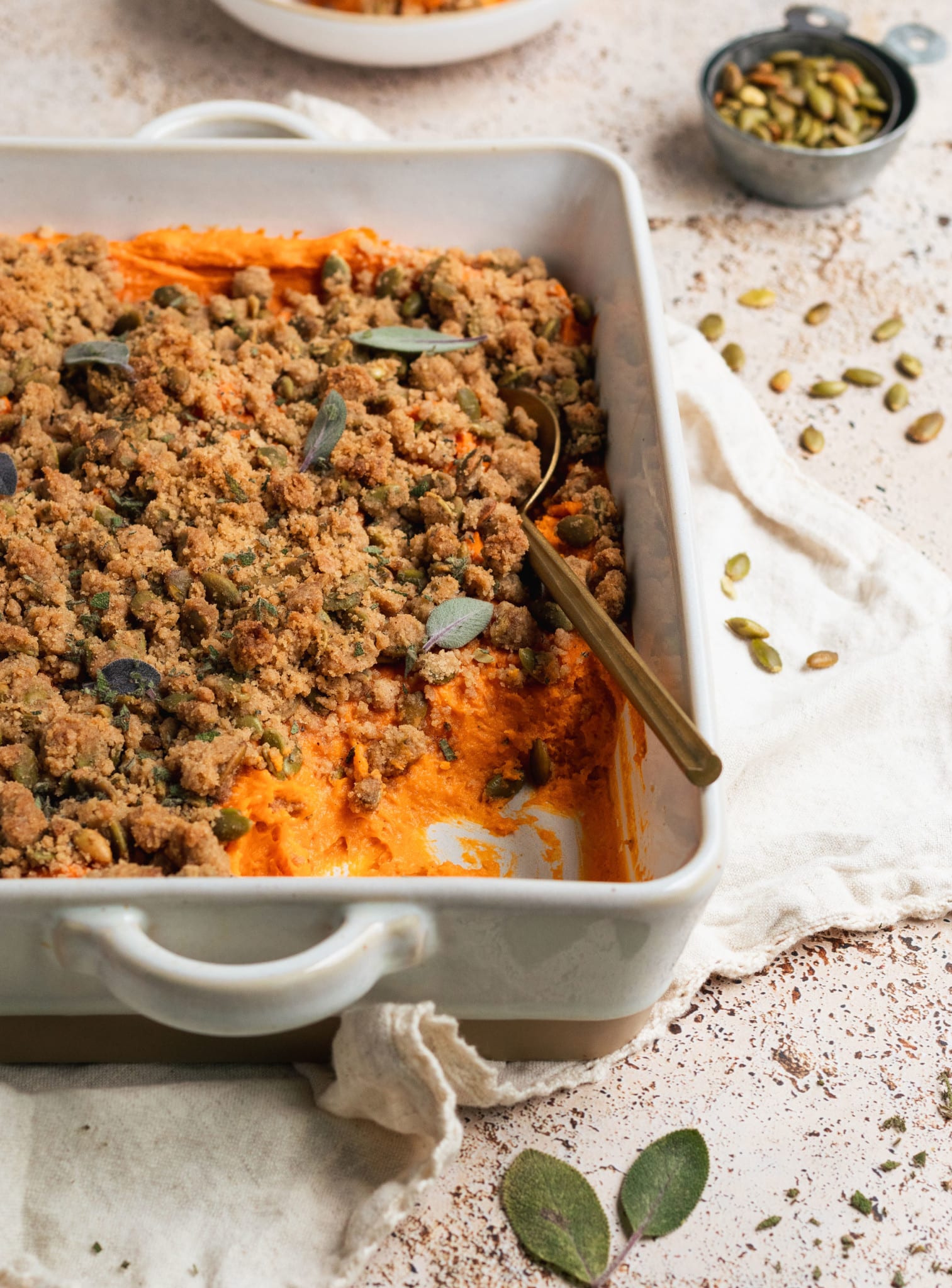 sweet potatoes in a white casserole dish topped with a brown sugar pepitas topping and fresh sage. 