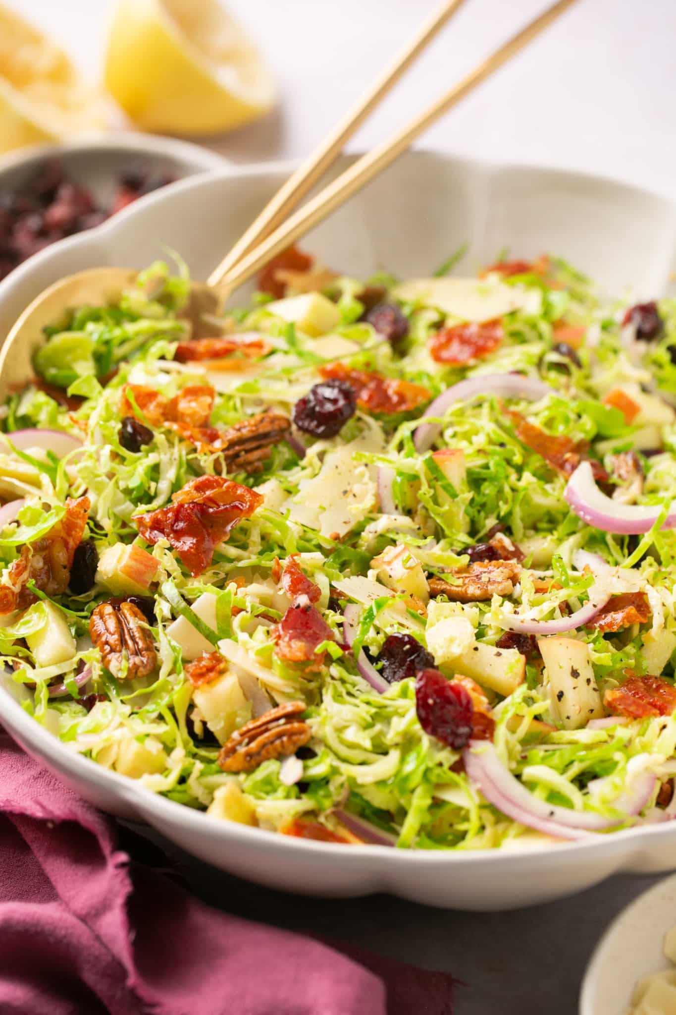 a large white bowl filled with brussels sprouts, cheese, onion, apple, dried cranberries, prosciutto, parmesan and salad dressing. 