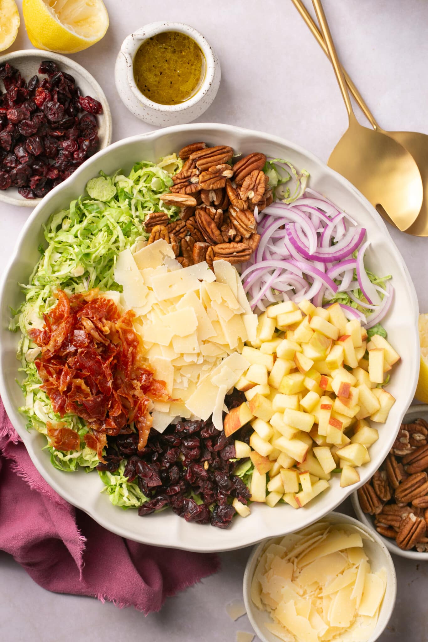 a large white bowl filled with brussels sprouts, cheese, onion, apple, dried cranberries, prosciutto, parmesan and salad dressing on the side. 