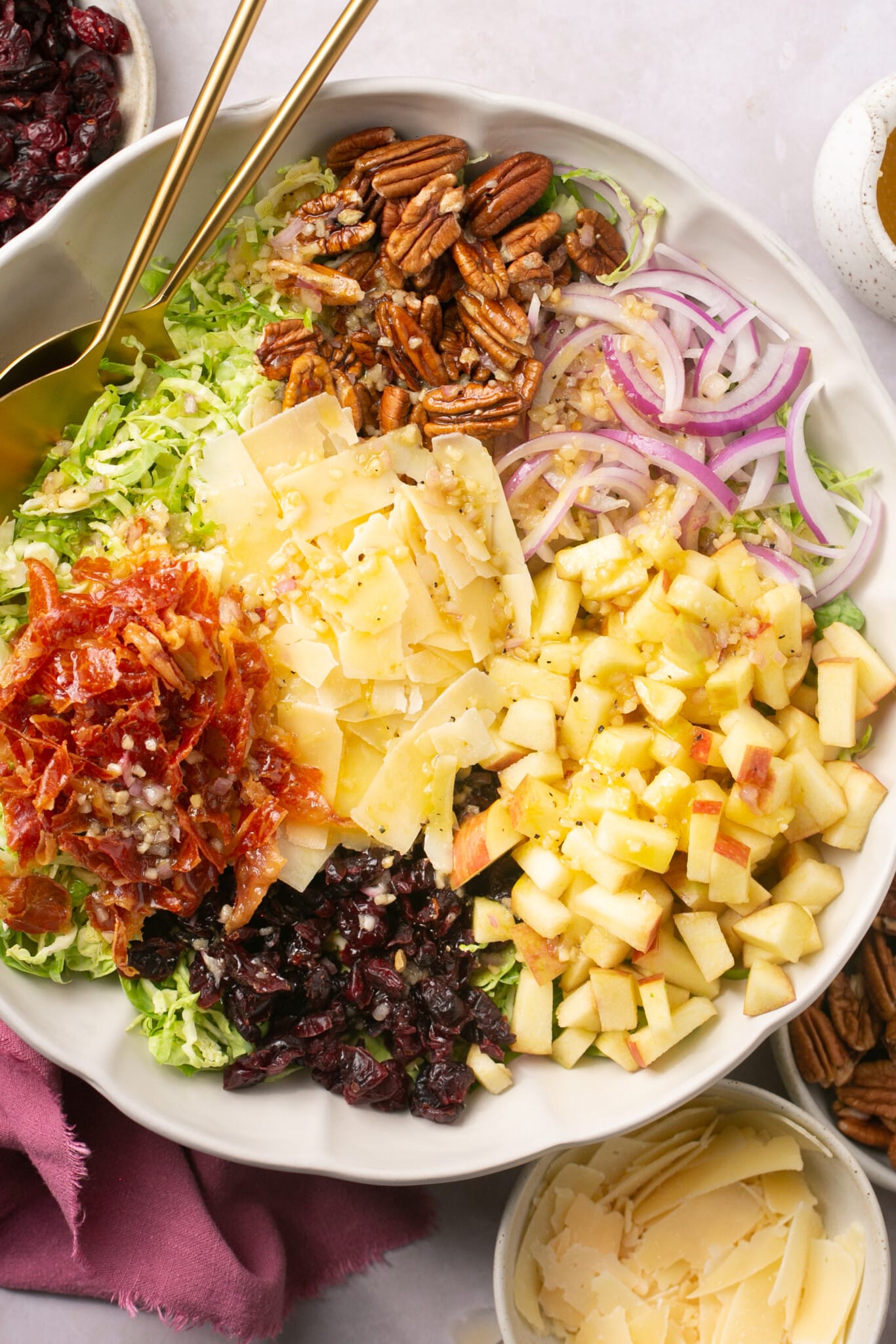 a large white bowl filled with brussels sprouts, cheese, onion, apple, dried cranberries, prosciutto, parmesan and salad dressing. 