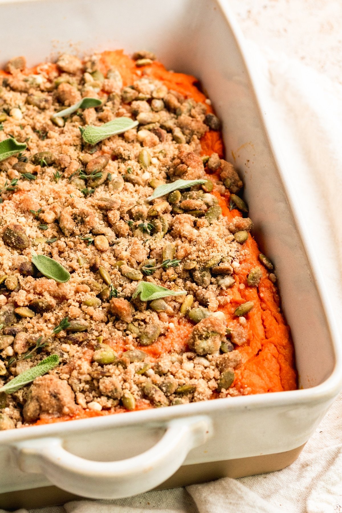 sweet potatoes in a white casserole dish topped with a brown sugar pepitas topping and fresh sage and fresh thyme. 