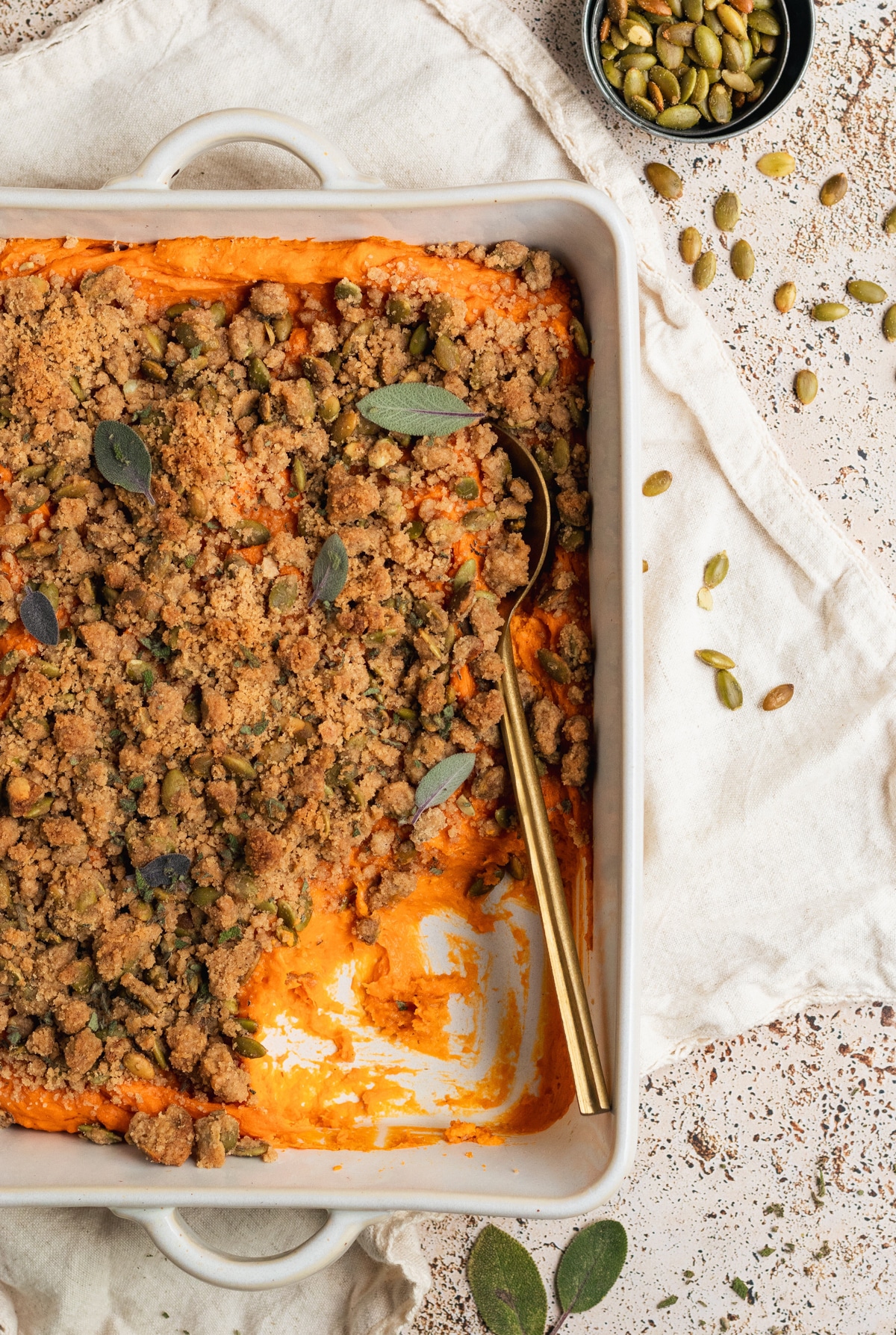 sweet potatoes in a white casserole dish topped with a brown sugar pepitas topping and fresh sage and fresh thyme with a gold spoon. 