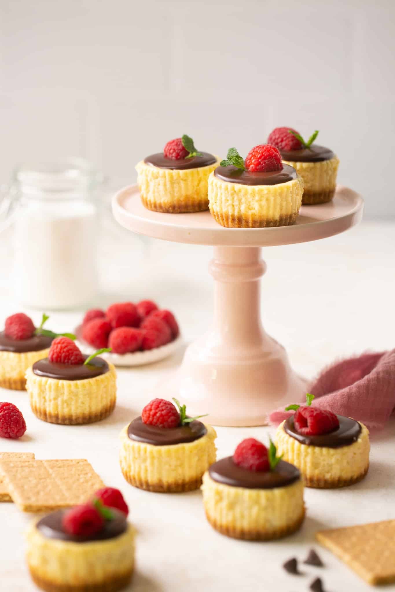 mini cheesecakes on a cake stand garnished with ganache, fresh raspberries and mint. 