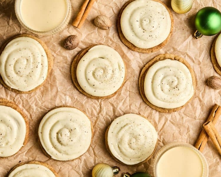 Cookies with swirl of white frosting on top of brown parchment paper surrounded by various christmas ornaments, sticks of cinnamon, and whole nutmeg.