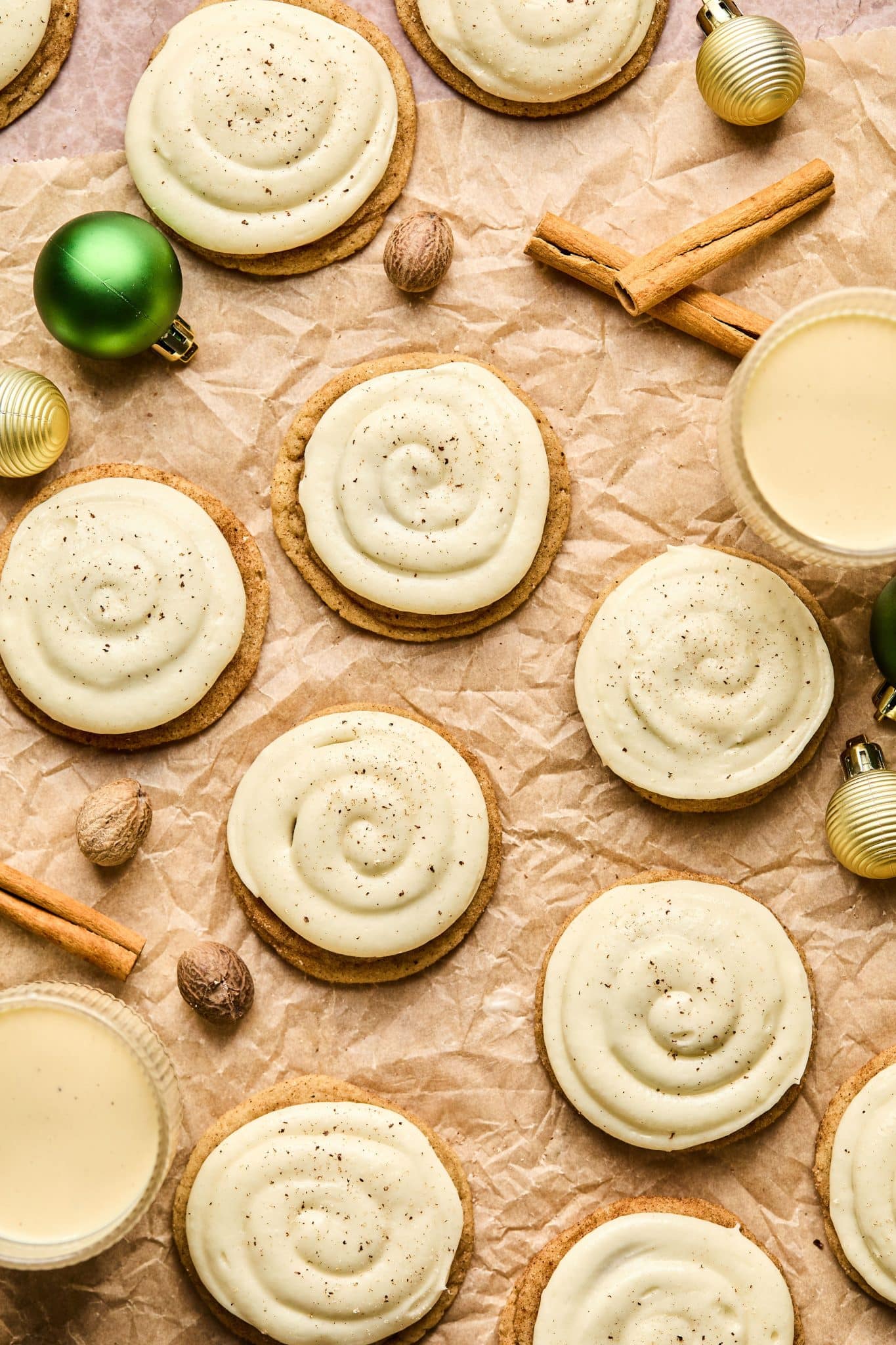 Cookies with white swirled frosting on brown parchment paper surrounded by various christmas ornaments, sticks of cinnamon, and whole nutmeg.