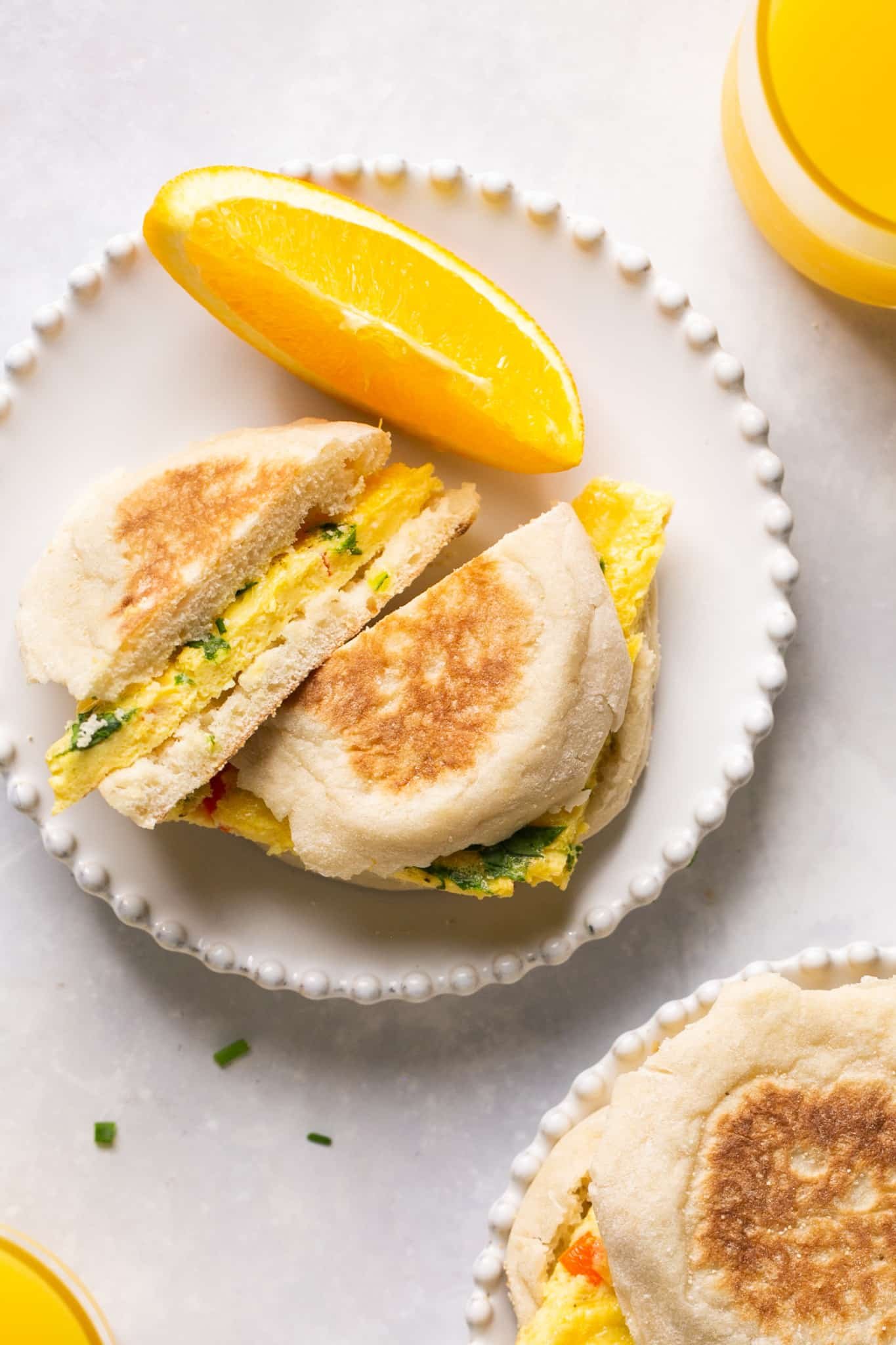 a breakfast sandwich on a white plate made with eggs on an english muffin and an orange slice on the side. 