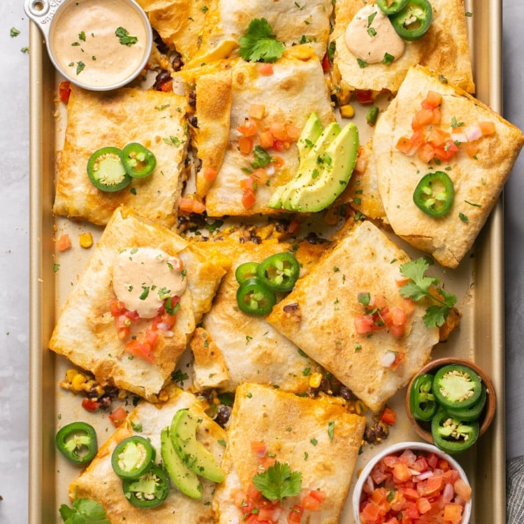 sheet pan with quesadillas cut into squares topped with avocado and jalapeno with two bowls of sauce and salsa