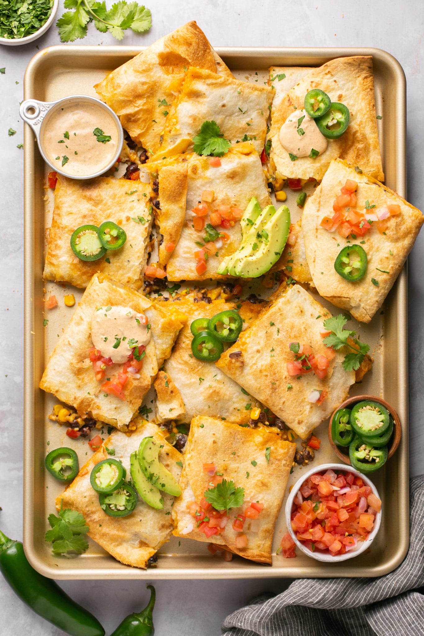 sheet pan with quesadillas cut into squares topped with avocado and jalapeno with two bowls of sauce and salsa