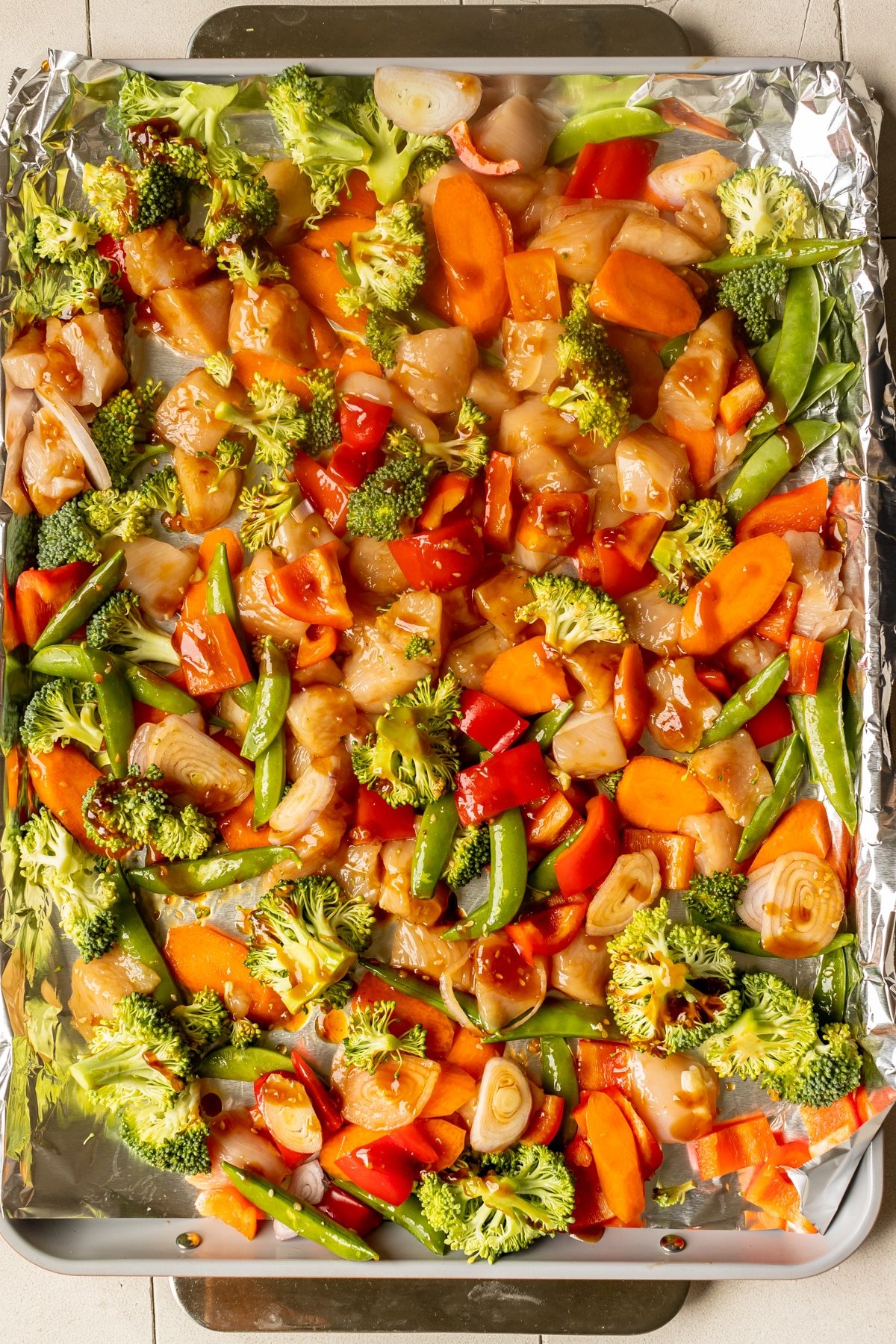 raw chicken and vegetables on a sheet pan lined with foil with sauce drizzled over the top. 