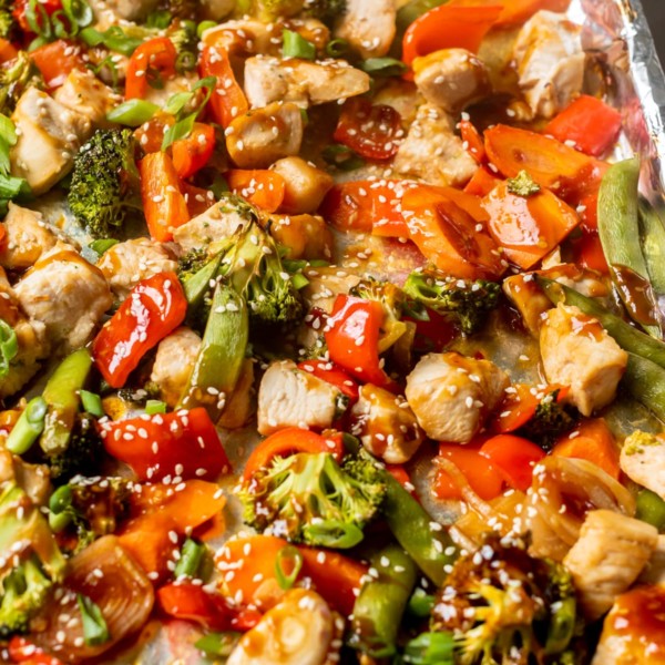sheet pan covered in foil with cooked chicken cubes and roasted veggies drizzled in sauce and topped with sesame seeds