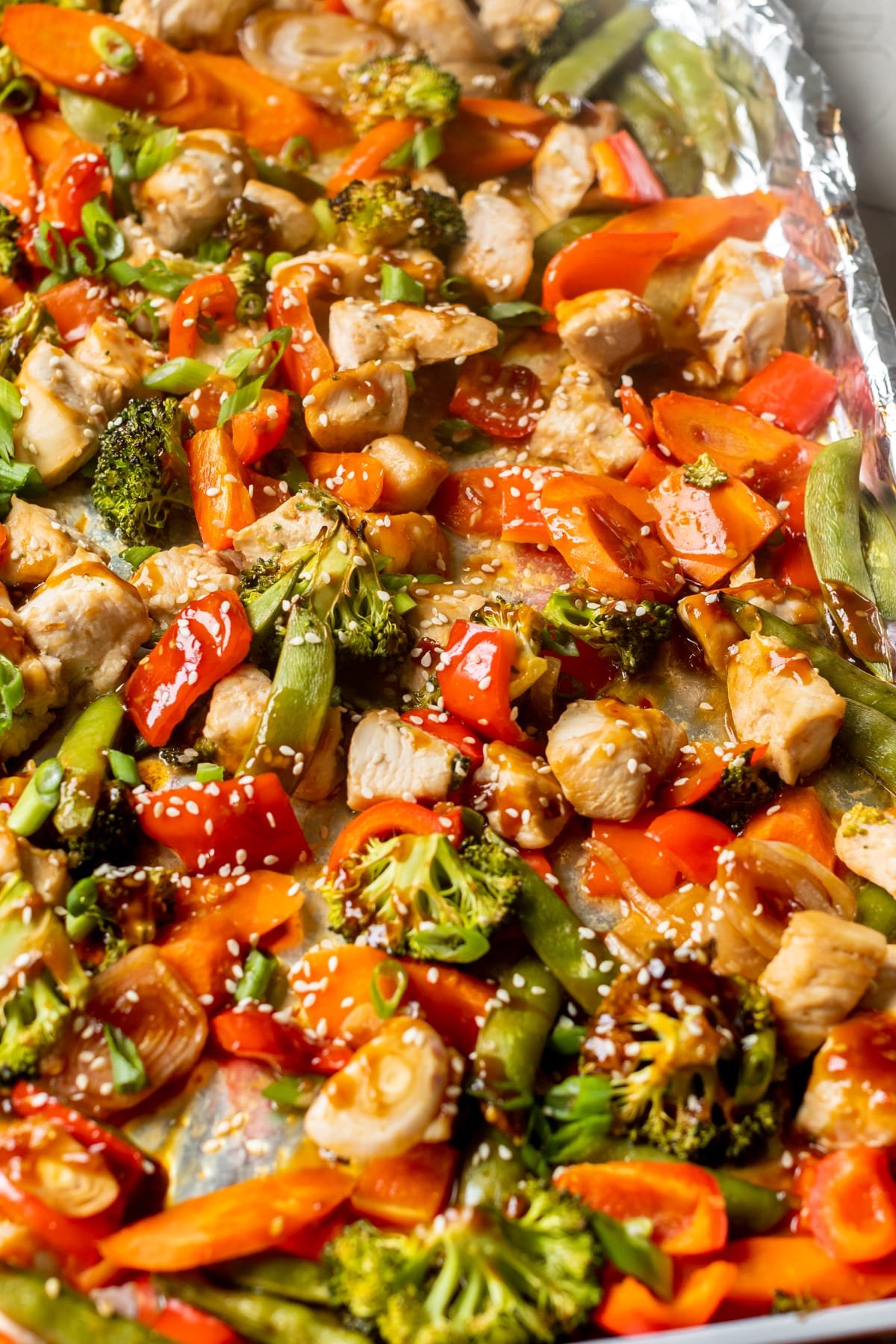 sheet pan covered in foil with cooked chicken cubes and roasted veggies drizzled in sauce and topped with sesame seeds