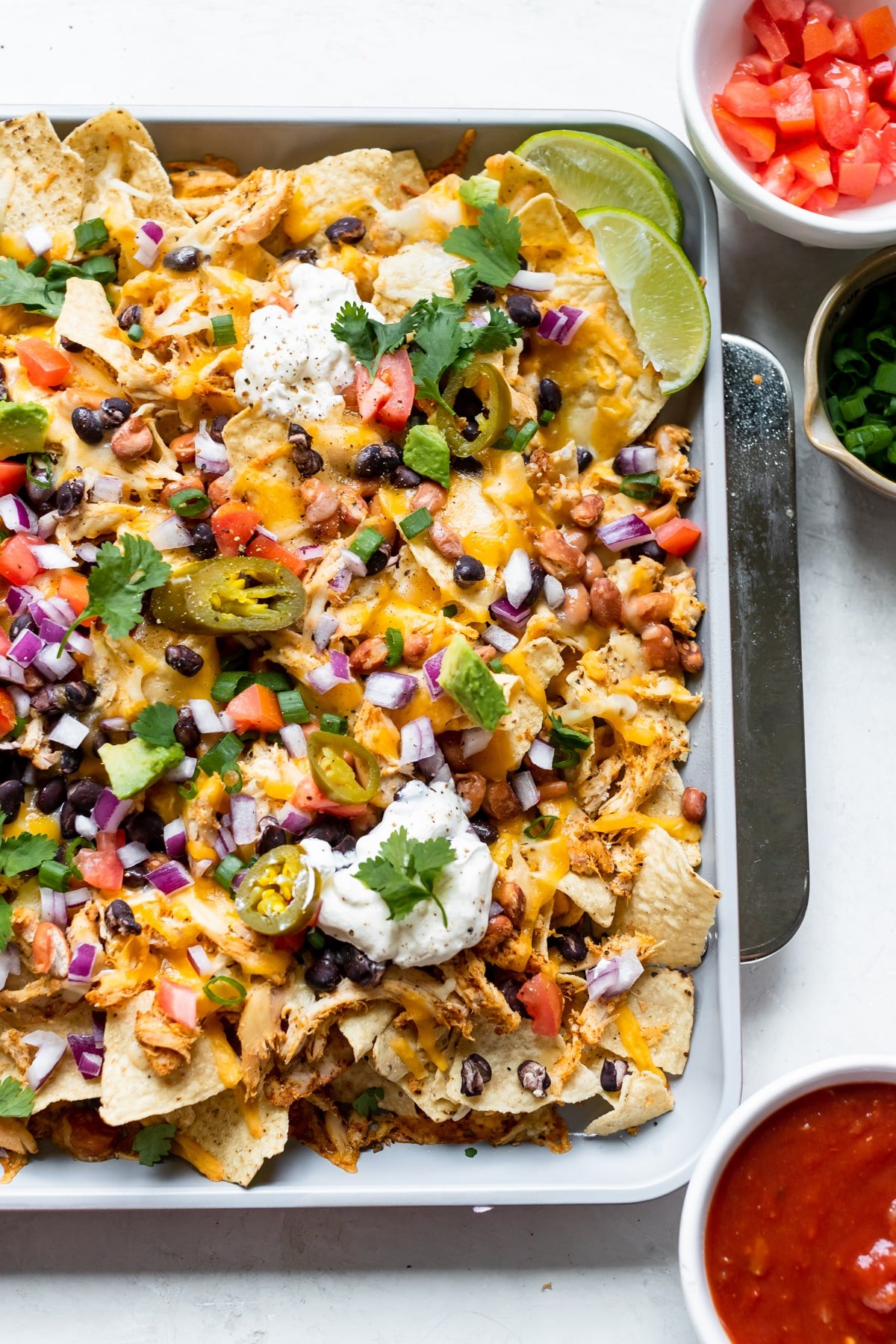 nachos on a a sheet pan garnished with green onions, jalapenos, sour cream, cilantro, red onions and tomatoes. 