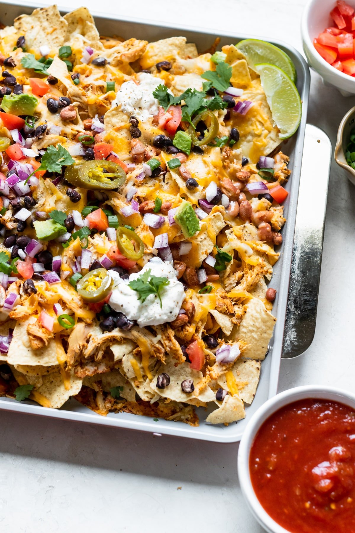 nachos on a a sheet pan garnished with green onions, jalapenos, sour cream, cilantro, red onions and tomatoes. 