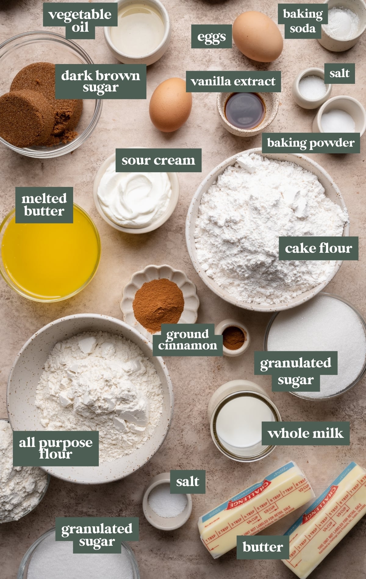 Overhead view of baking ingredients arranged on a light countertop, each labeled, including cake flour, all-purpose flour, granulated sugar, dark brown sugar, eggs, sour cream, whole milk, vanilla extract, vegetable oil, melted butter, butter sticks, ground cinnamon, baking powder, baking soda, and salt in small bowls and ramekins.